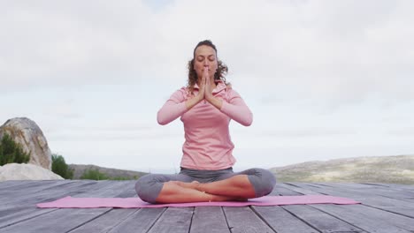 Caucasian-woman-practicing-yoga-meditation-in-lotus-position-on-deck-in-rural-mountainside-setting