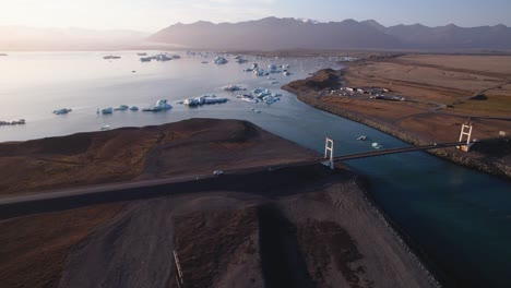 Serene-glacial-lagoon-with-icebergs,-a-bridge-crossing,-and-a-backdrop-of-mountains-at-dusk-from-aerial-perspective-in-Iceland