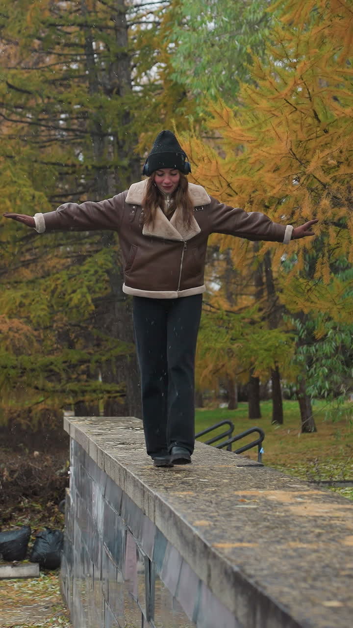 In drizzling rain, smiling girl wearing headphone, black knit cap, brown shearling jacket, black trousers balances gracefully with arms outstretched while walking joyfully on wide stone surface