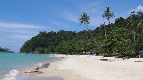 Imágenes-En-Cámara-Lenta-De-Olas-Que-Bañan-A-Dos-Mujeres-Tomando-El-Sol-En-Una-Costa-Tropical