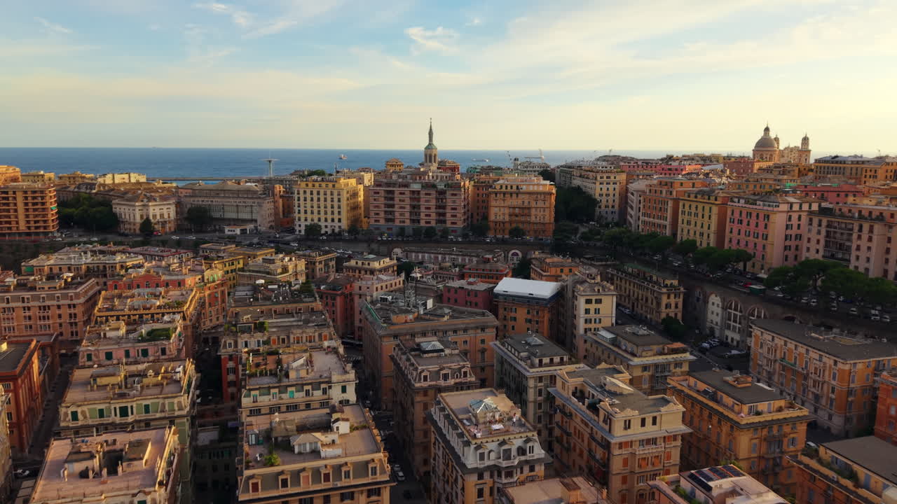 Drone flies forward above the rooftops of Genoa city center at sunset, showing colorful buildings, symmetrical streets, a church tower, the sea, and the sky in the background