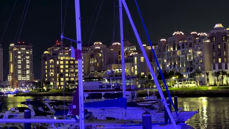 Primer-Plano-Yate-En-El-Canal-De-Agua-De-Mar-Del-Golfo-árabe-Con-La-Bandera-De-Qatar-En-Perla-Qanat-Quartier-De-Venecia-En-Doha-En-La-Noche-Espectáculo-Fondo-Oscuro-De-Palmeras-Edificios-Torre-Del-Hotel-Refleja-La-Luz