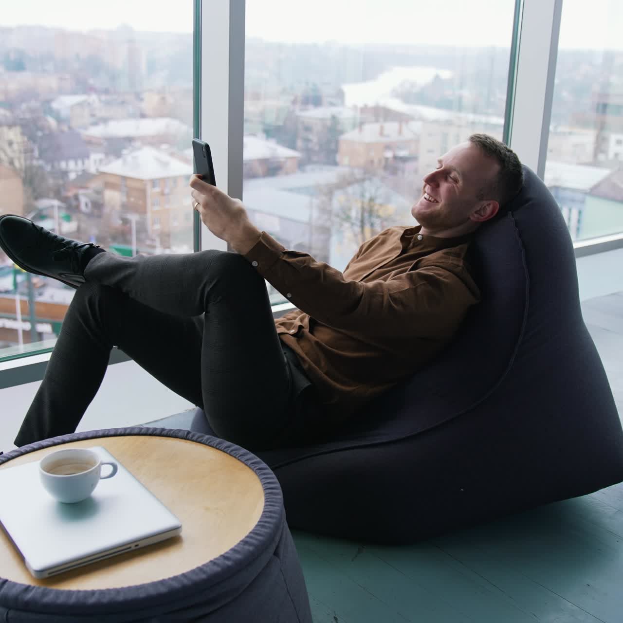 Young businessman relaxing near the window. Handsome man sitting in a cosy chair and having a video conversation with the city window view. Profile view