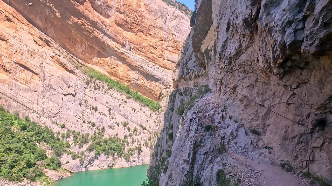A young woman carefully walking along the narrow path of Congost de Mont-Rebei, with steep cliffs and the turquoise river below, under a clear blue sky.