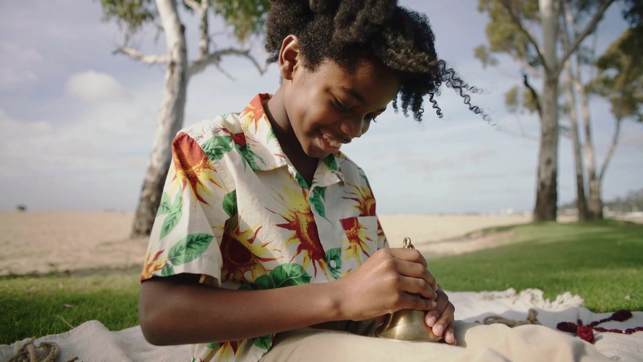 Campana che attira l'attenzione, un bambino con camicia floreale ispeziona una campana di ottone con batacchio su una coperta da spiaggia.