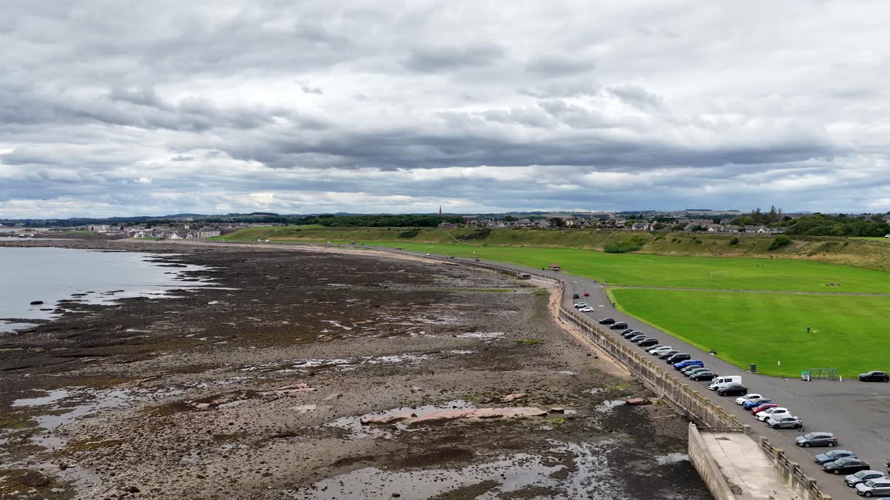 Drone pans above rocky shoreline, green park, parked cars, under cloudy daylight in Dundee, Scotland