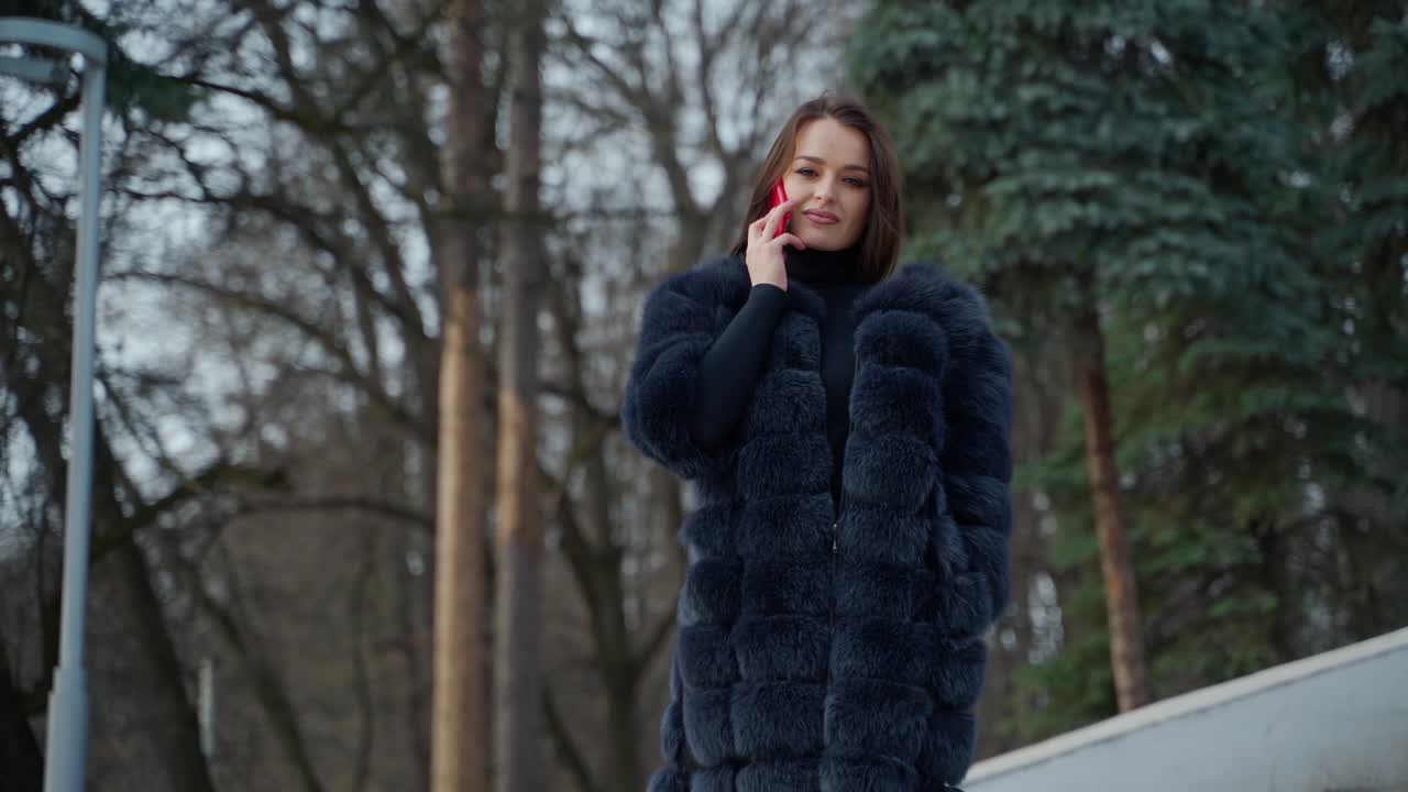 Beautiful model in warm coat on nature background. Portrait of a lovely girl in fur coat talking the red phone and looking at camera in winter park.