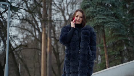 beautiful model in warm coat on nature background. portrait of a lovely girl in fur coat talking the red phone and looking at camera in winter park.