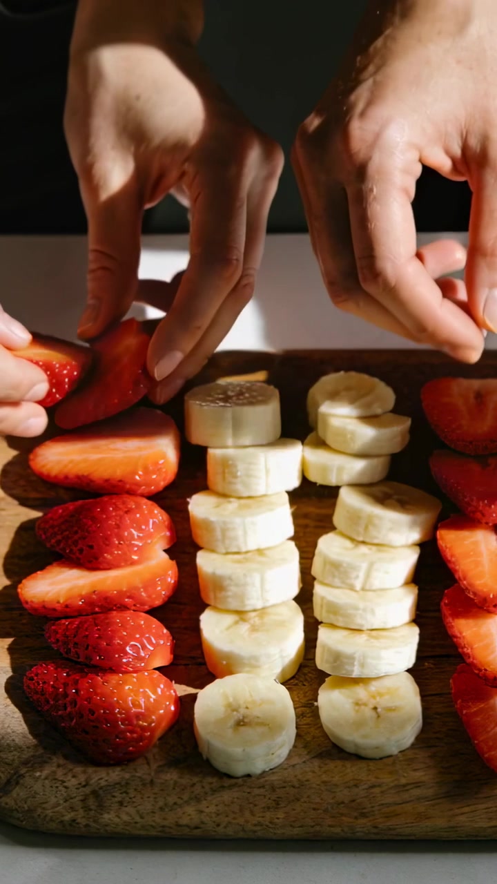 Video verticale: Mani che dispongono mezze fragole e banana su un tagliere in cucina per simmetria.