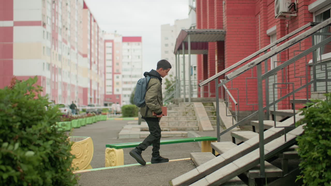 Back view of young boy with backpack walking outdoors holding book towards bench, blurred background of residential buildings and parked cars capturing daily routine