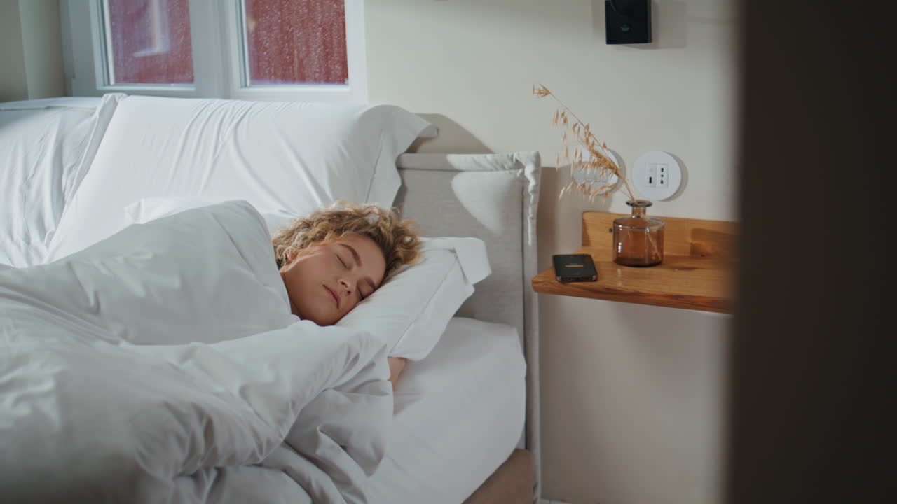 Serene woman sleeping home in morning sunlight. Smartphone alarm ringing table