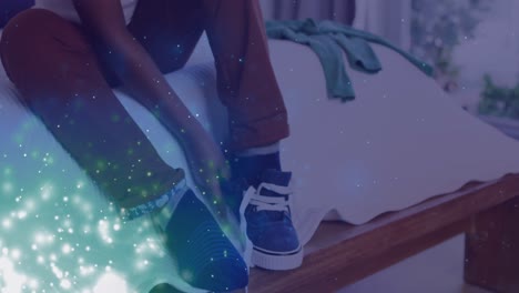 animation of lights over feet of african american boy tying shoe on bed