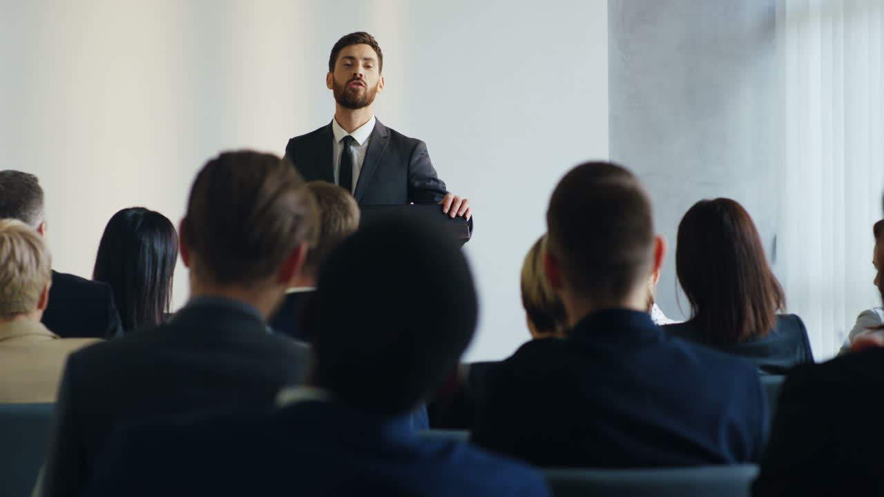 Caucasian businessman speaker on a podium wearing formal clothes and talking in a conference room in front to many people