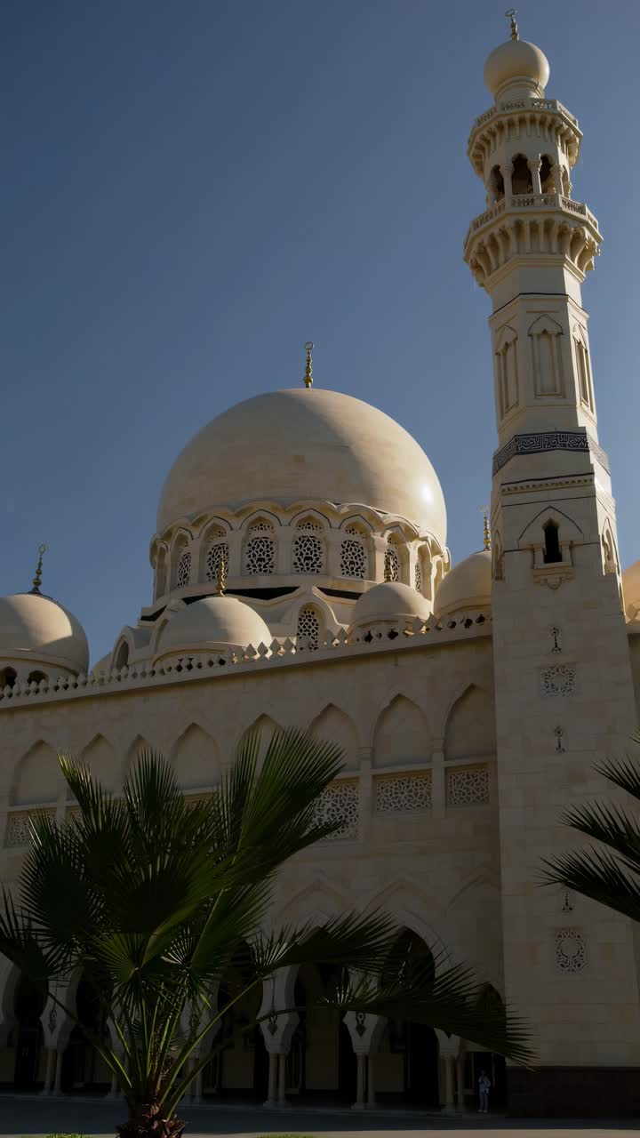 Low-angle video shot of a grand mosque with intricate domes and minaret against a clear blue sky