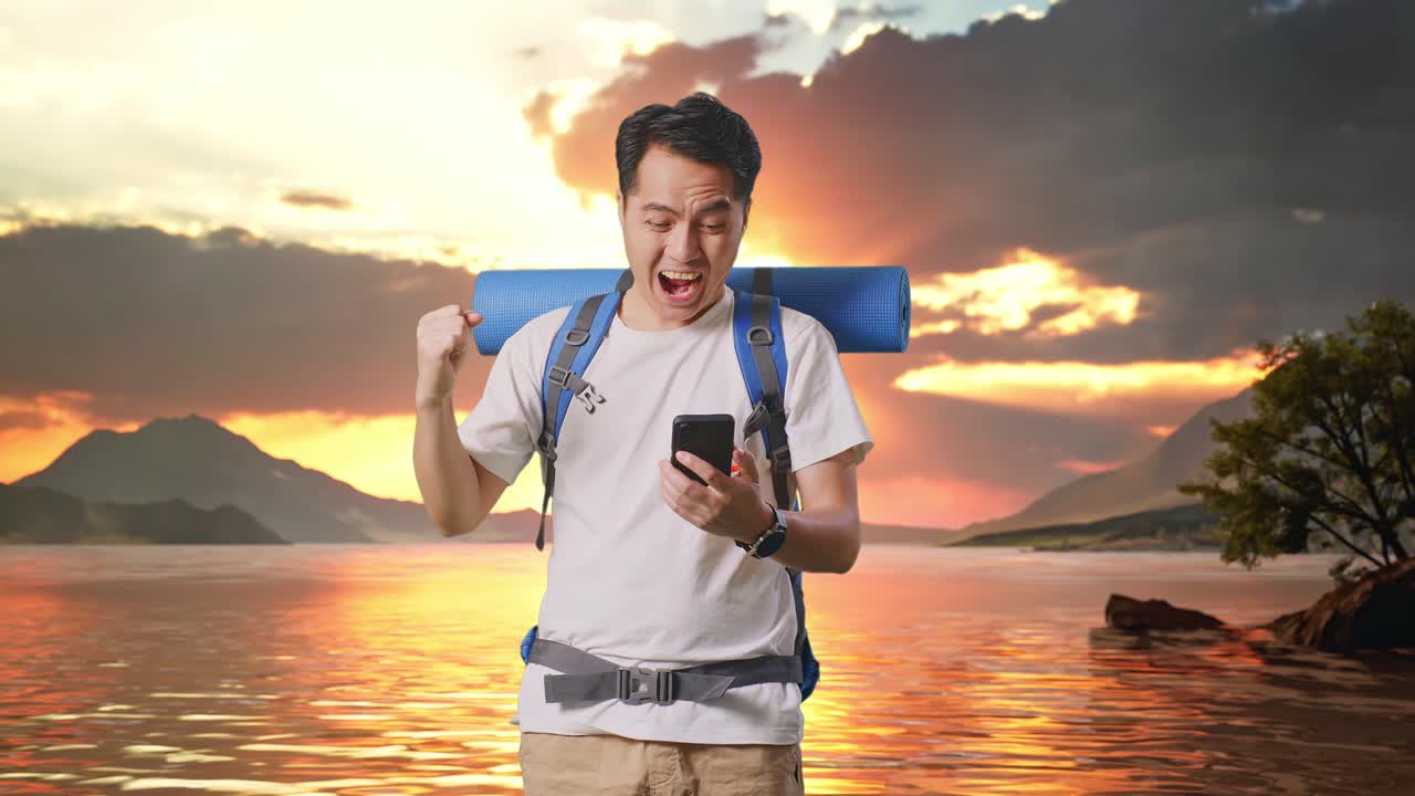 excursionista masculino asiático con mochila de montañismo mirando el teléfono inteligente y gritando meta celebrando el éxito en un lago