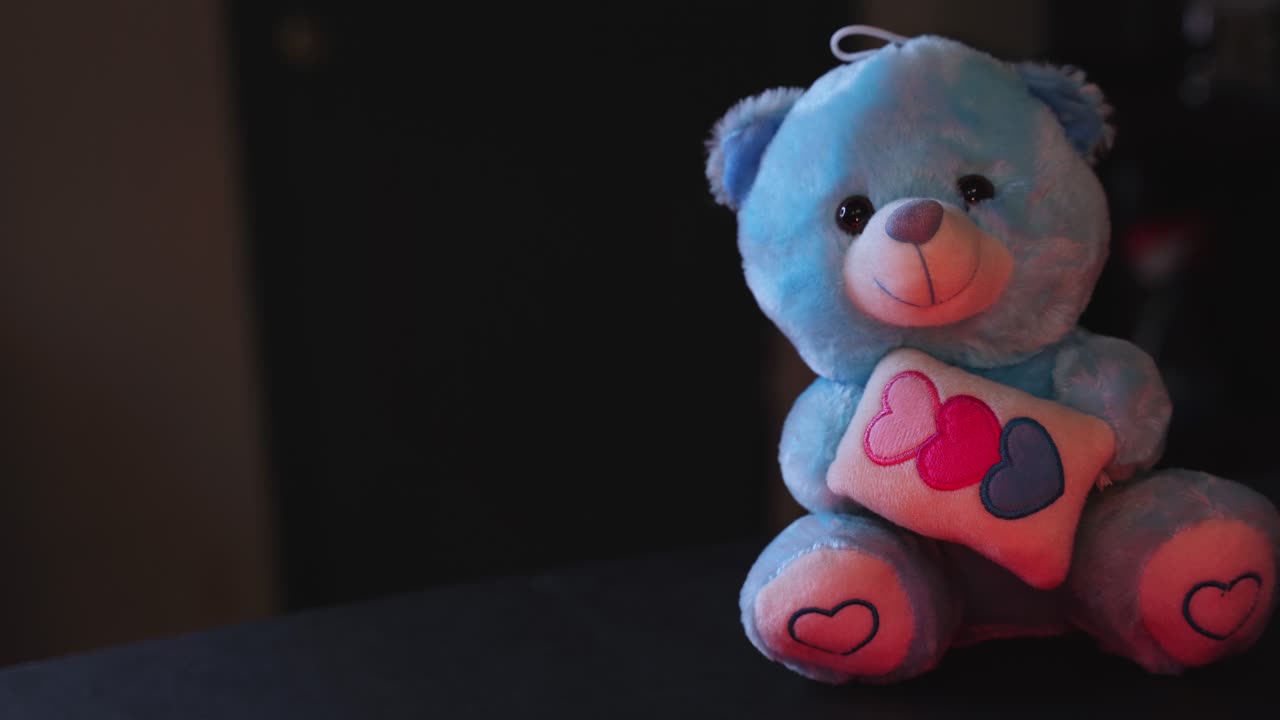 Close-up boneka beruang biru mewah memegang bantal hati, diterangi dengan cahaya aksen merah hangat dengan latar belakang dalam ruangan yang gelap, sempurna untuk tema Hari Valentine, romansa, dan dekorasi yang nyaman.