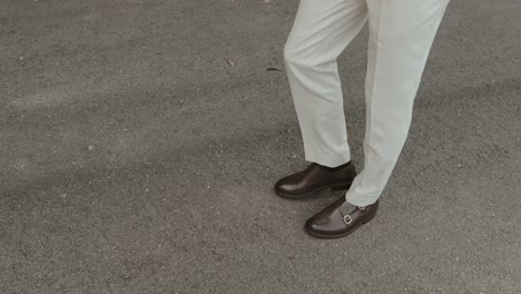 close up of groom walking in brown leather shoes and cream trousers on asphalt road
