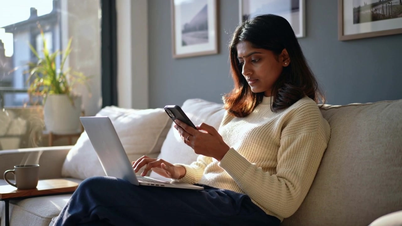 Alternating between smartphone and laptop, woman in cream sweater typing on sofa working at home