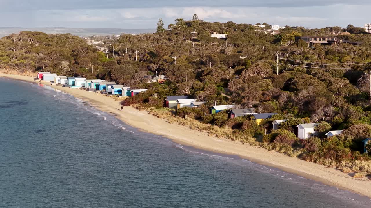 Drone glides above sandy shoreline, colorful bathing boxes, and coastal vegetation in natural daylight