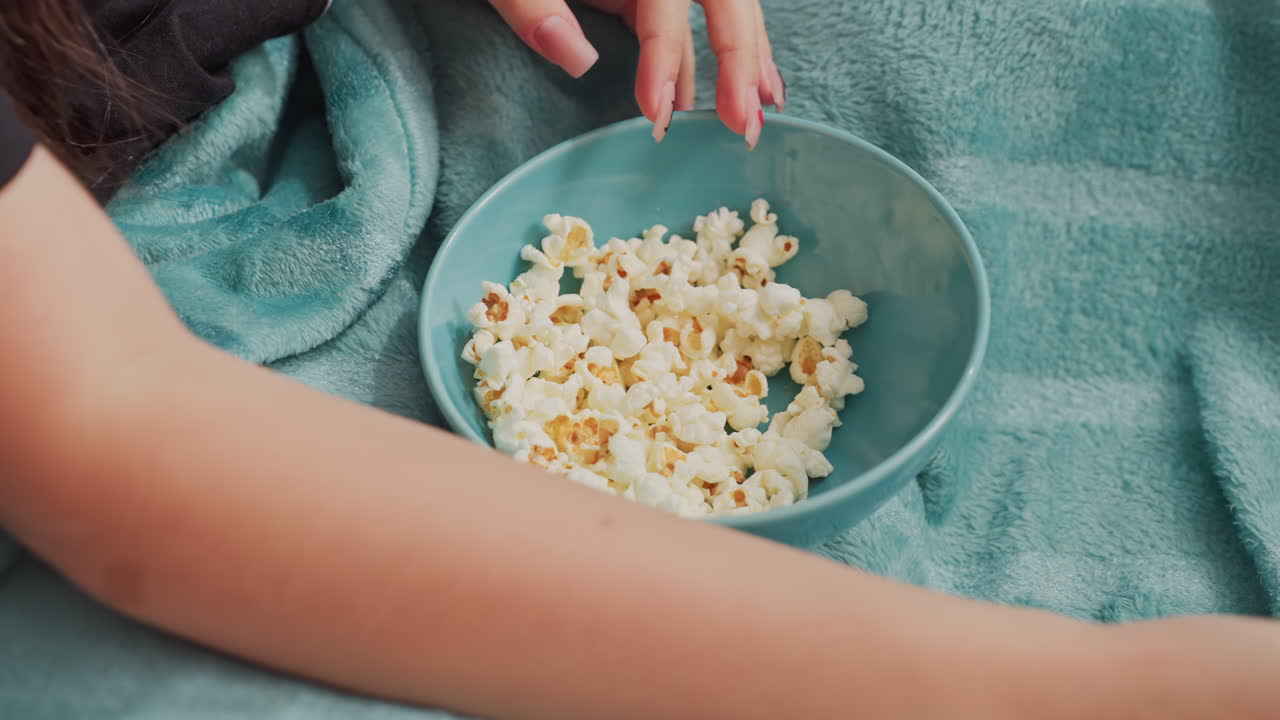 Close up of hands reaching for popcorn from blue bowl placed on teal blanket, scene captures relaxed indoor moment of casual snacking, leisure
