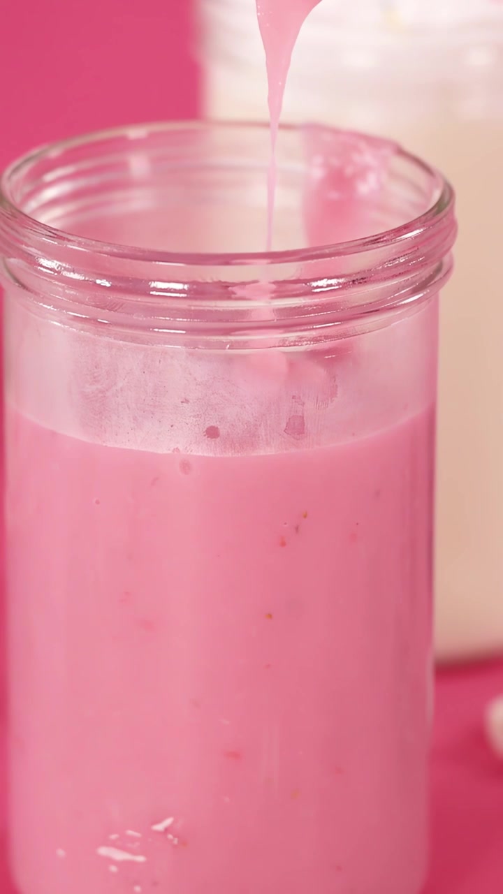 Un milkshake alla fragola si versa dolcemente in un barattolo di vetro trasparente, con uno sfondo rosa vivace e un'illuminazione brillante e uniforme.