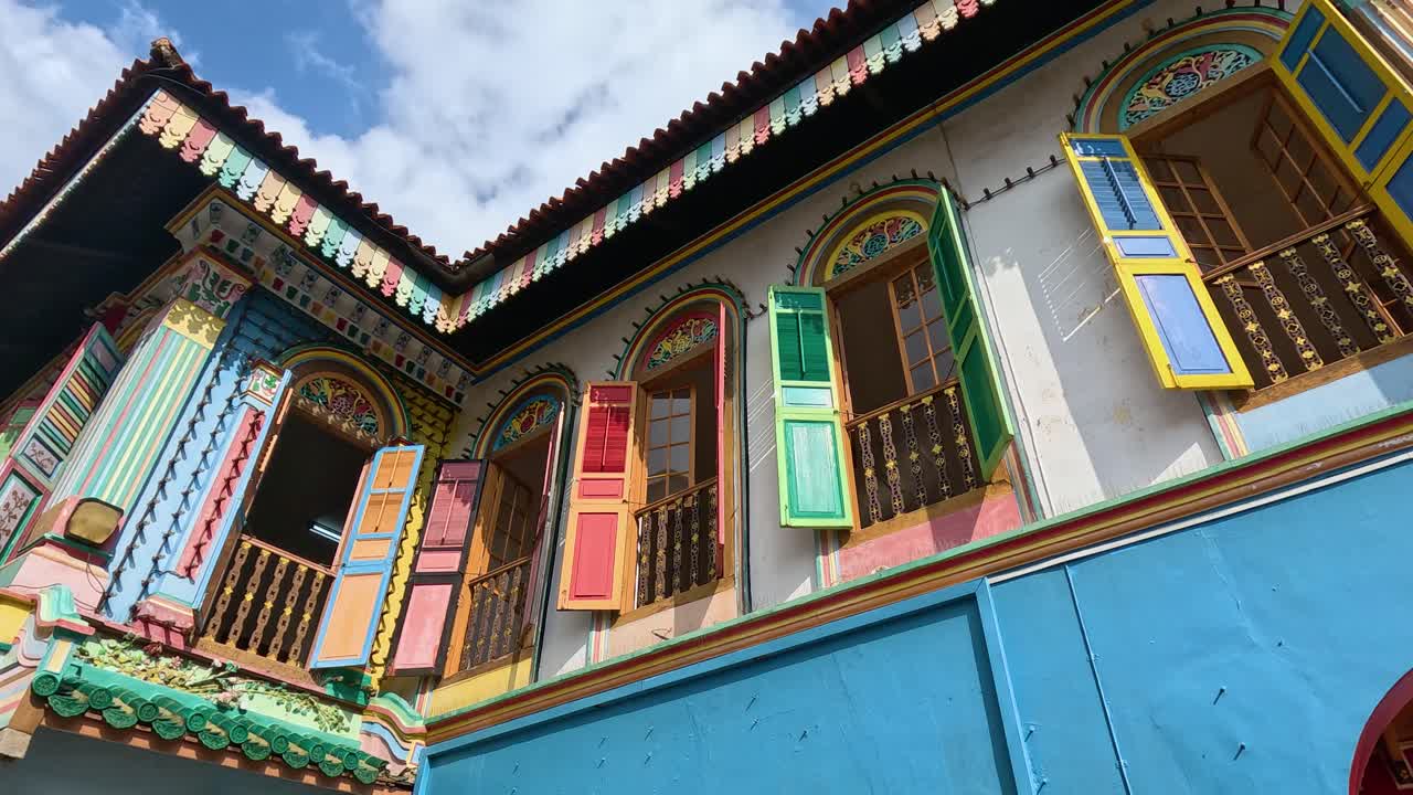 Camera tilts down a vibrant, historic shophouse with ornate windows and bright blue walls under daylight, revealing a narrow urban alley in Singapore