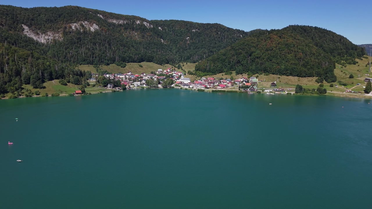 Een luchtfoto toont een schilderachtig dorpje genesteld aan de voet van beboste bergen naast een uitgestrekt, blauw meer op een heldere dag.