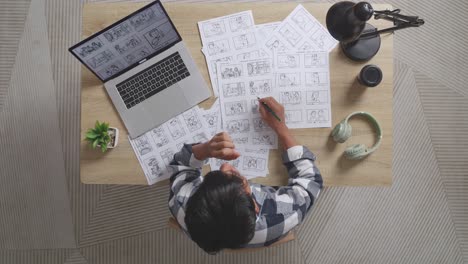 vista superior de un artista masculino asiático cansado bostezando y durmiendo mientras dibuja un guión gráfico en la mesa con una computadora portátil y auriculares en el estudio