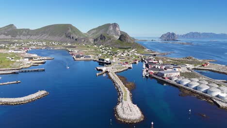 Aerial view of Værøy harbor: colorful village by the sea, dramatic mountains rising behind, and the endless blue of the Norwegian coast