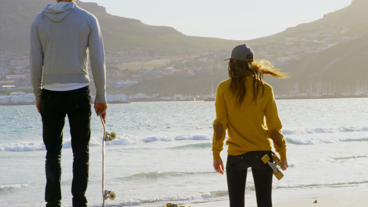 해변에 서서 바다를 바라보는 젊은 백인 스케이트보더의 뒷면 4k