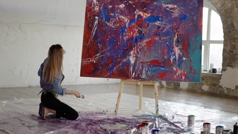 woman uses wide brush and color paints splashing gesture on canvas standing on knees