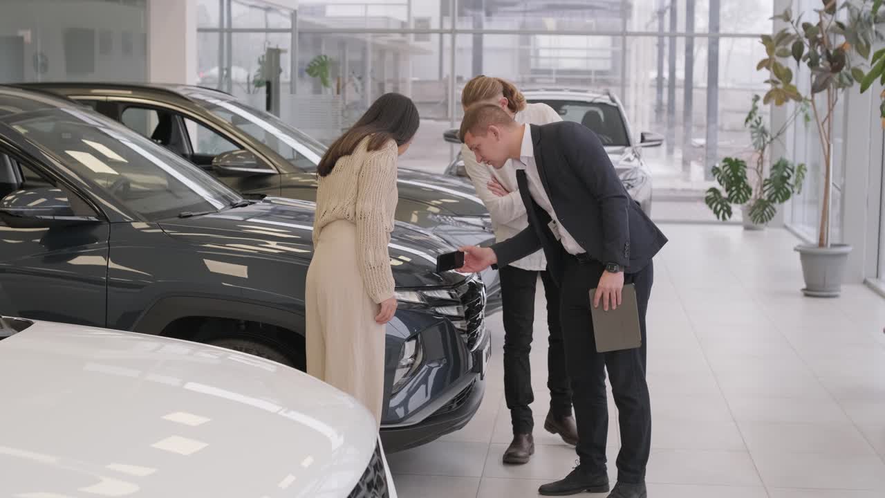 un joven vendedor de coches mostrando a una pareja de jóvenes un nuevo automóvil en el salón de concesionarios.