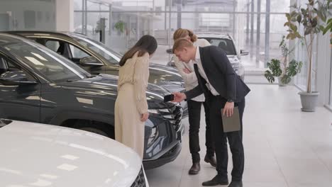 young car salesman showing to young couple new automobile at dealership salon.