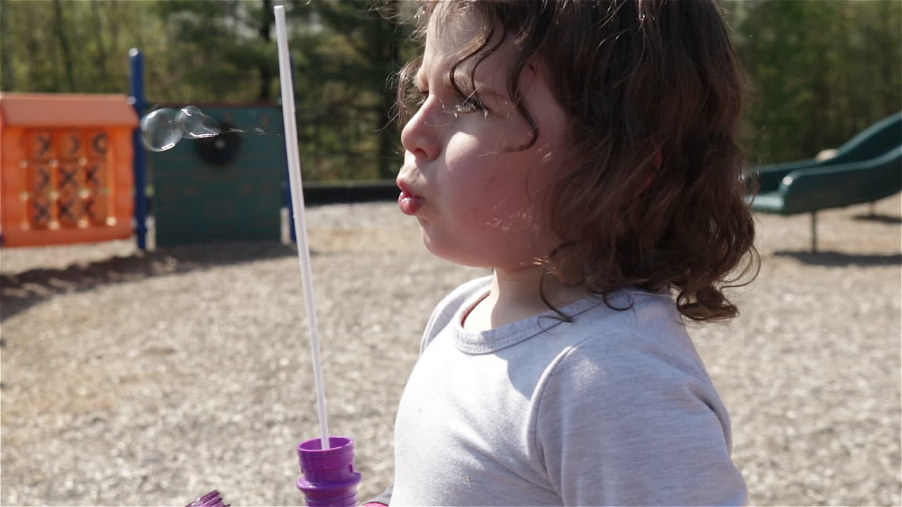 Young girl blows bubbles with wand at playground, slow motion close up