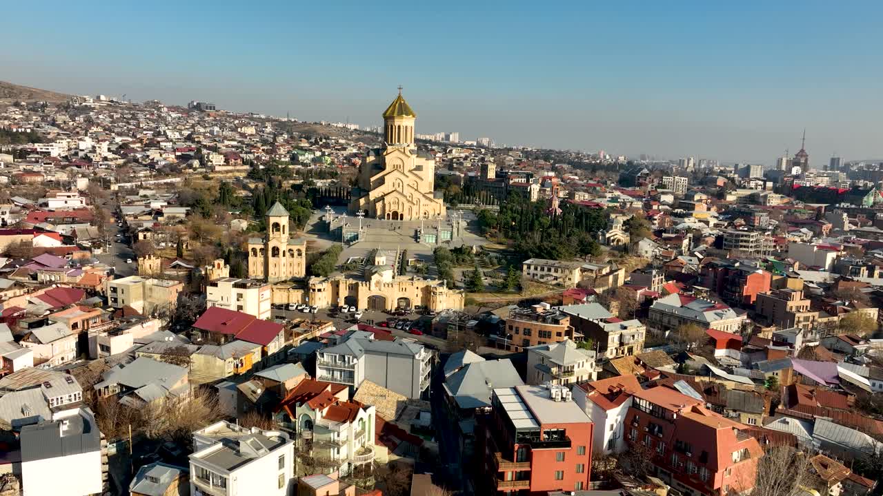 Foto de drone mostrando uma vista ampla de Tbilisi com a Catedral de Sameba se elevando acima das áreas residenciais. A foto destaca a estrutura urbana da cidade e o marco religioso icônico.