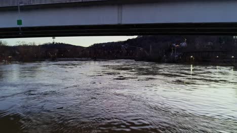 Drohne-Fliegt-Unter-Brücke,-Während-Das-Auto-In-Einem-Kalten-Winter-Bei-Sonnenuntergang-über-Einen-Fluss-Fährt