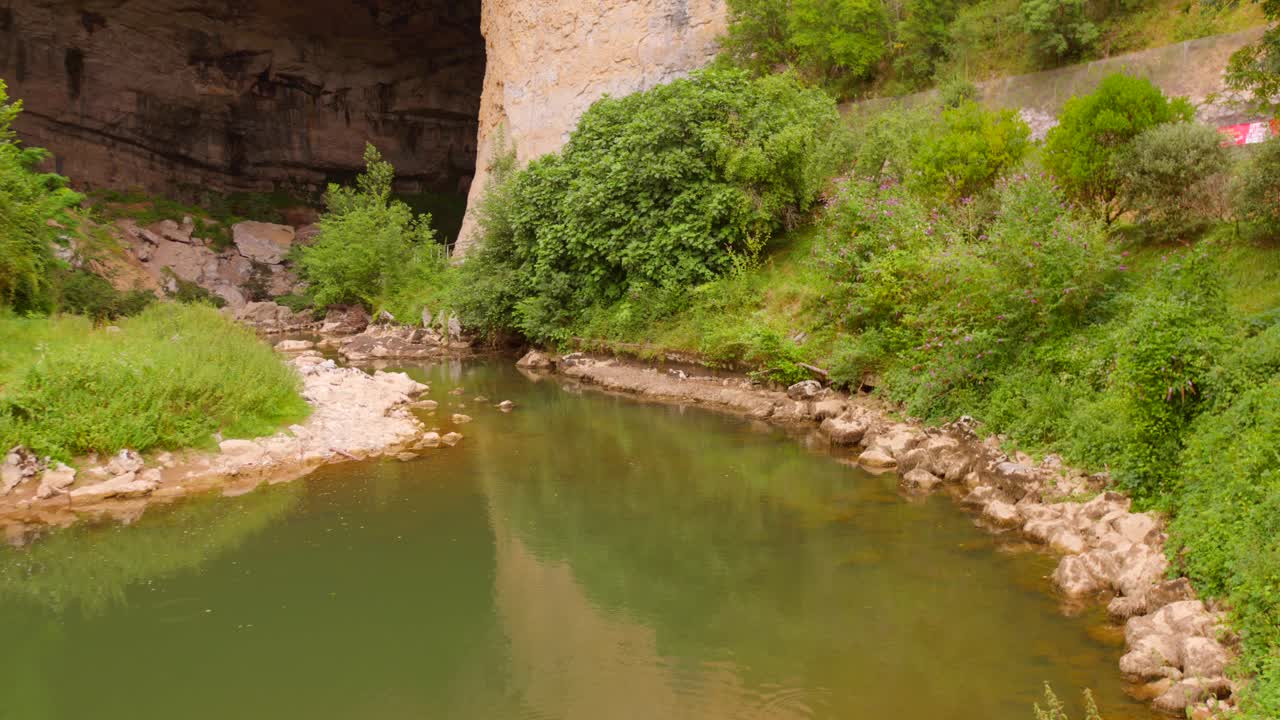 View of a small river flowing near the entrance of Grotte du Mas d’Azil, France, with limestone cliffs and green vegetation