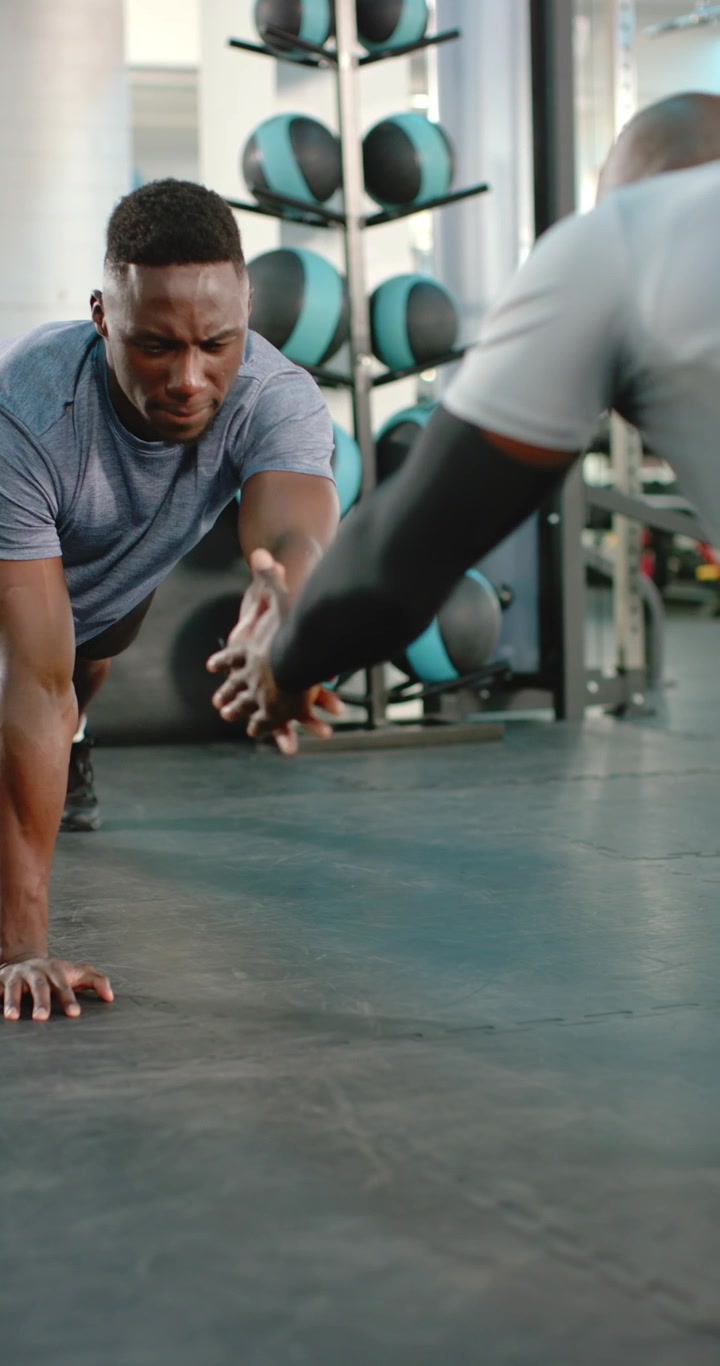Vídeo vertical: Dupla masculina afro-americana fazendo prancha e descendo para flexões com toque na academia para a saúde.