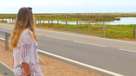 Female-Traveler-Seated-On-A-Wooden-Fence-At-The-Road-In-Ebro-Delta-Park,-Spain