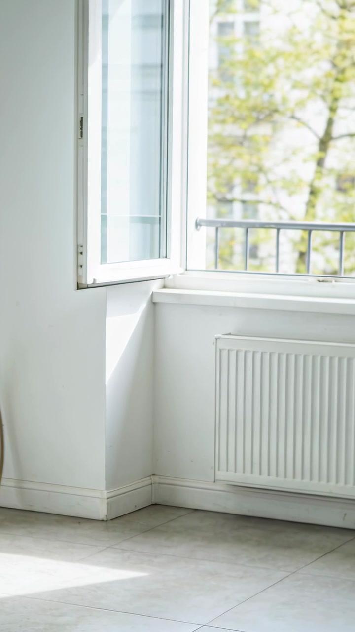 Vertical video: Entering small blue robot vacuum cleaning floor near open window, with radiator