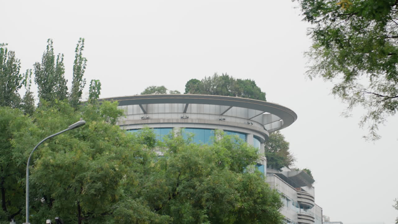 Moderno edificio rotondo che spunta tra gli alberi, facciata in vetro blu e balcone curvo visibili sopra la chioma frondosa sotto un cielo coperto, accenni a una terrazza panoramica e un giardino, tranquillo lampione in primo piano, calma