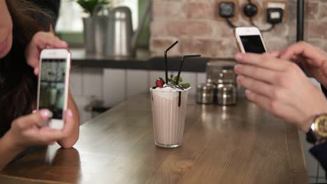 Young-man-and-woman-taking-picture-of-a-glass-with-milk-shake-with-cherry-on-the-top-and-straw-on-a-wooden-table-in-cafe