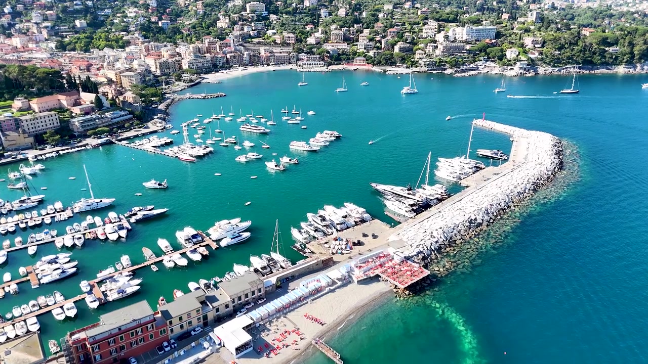 Prospettiva aerea soleggiata di barche ormeggiate in acque turchesi lungo la costa della Riviera Italiana.