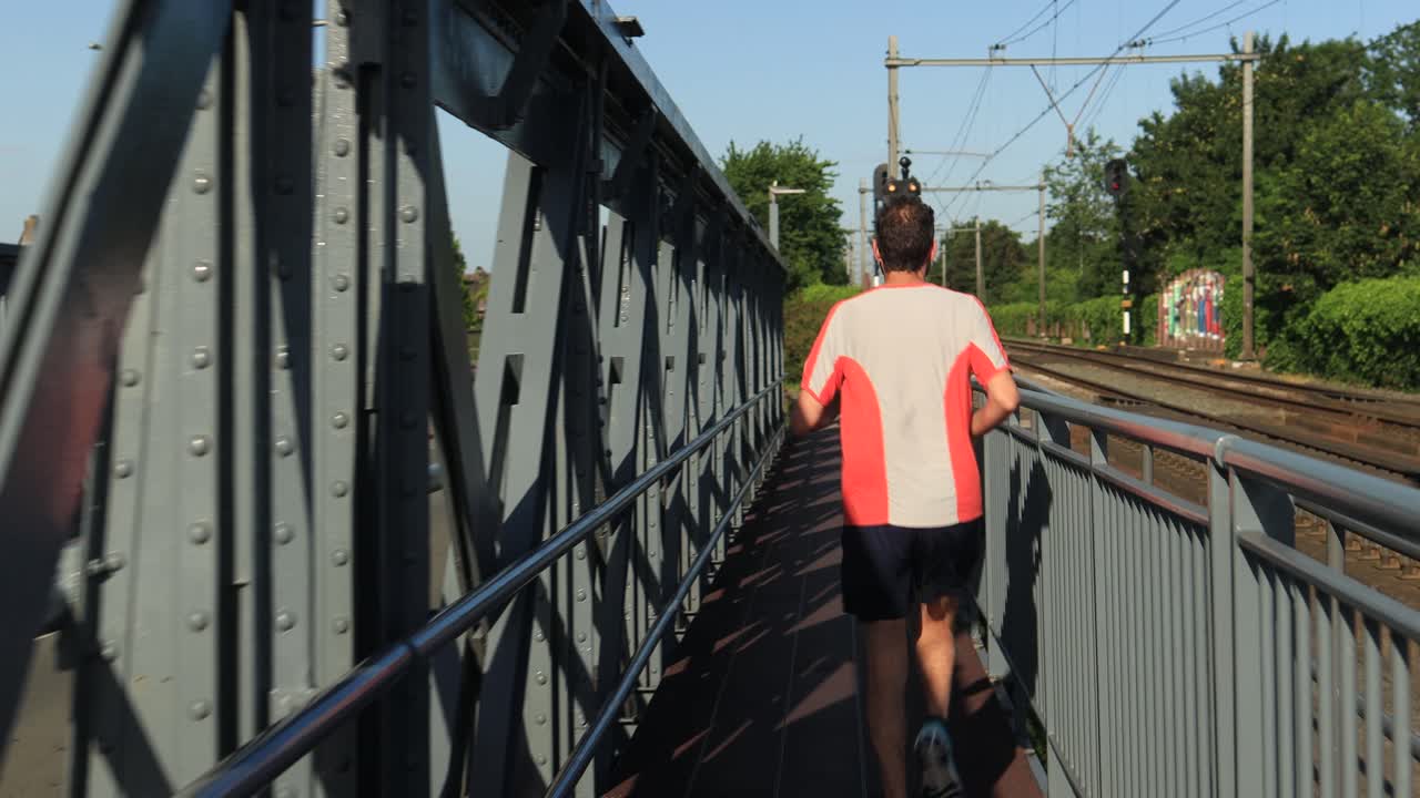 cadans de carrera constante de un corredor de rastro masculino visto desde atrás seguido por un puente de acero a lo largo de las vías del tren