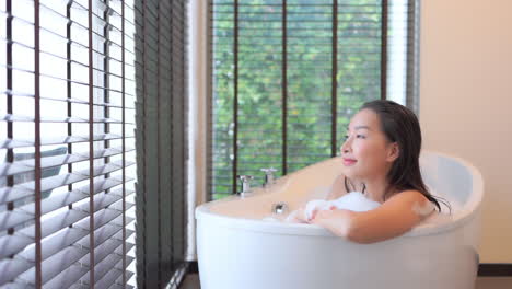 Static slow-motion shot of a sexy Asian lady relaxing in a bubble bath in a white tub while looking at the view outside the window