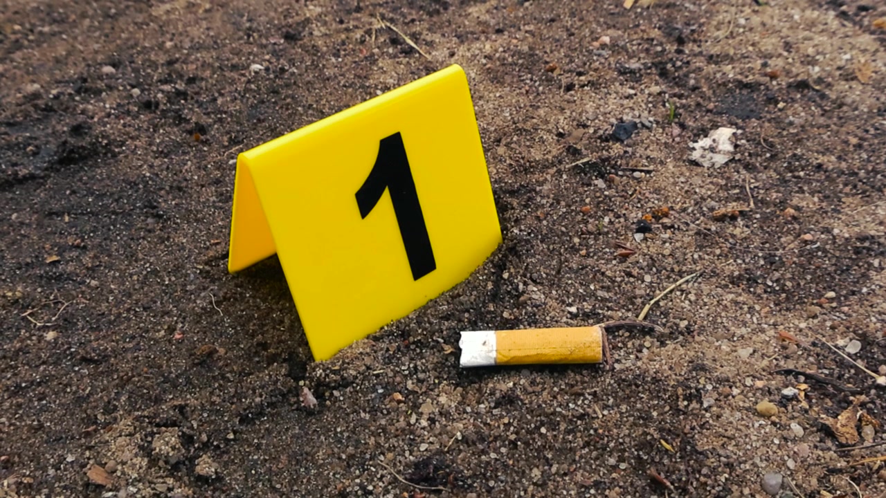 Marcatore di prova giallo con il numero uno accanto a un mozzicone di sigaretta a terra durante un'indagine forense su una scena del crimine all'aperto, evidenziando potenziali prove di DNA