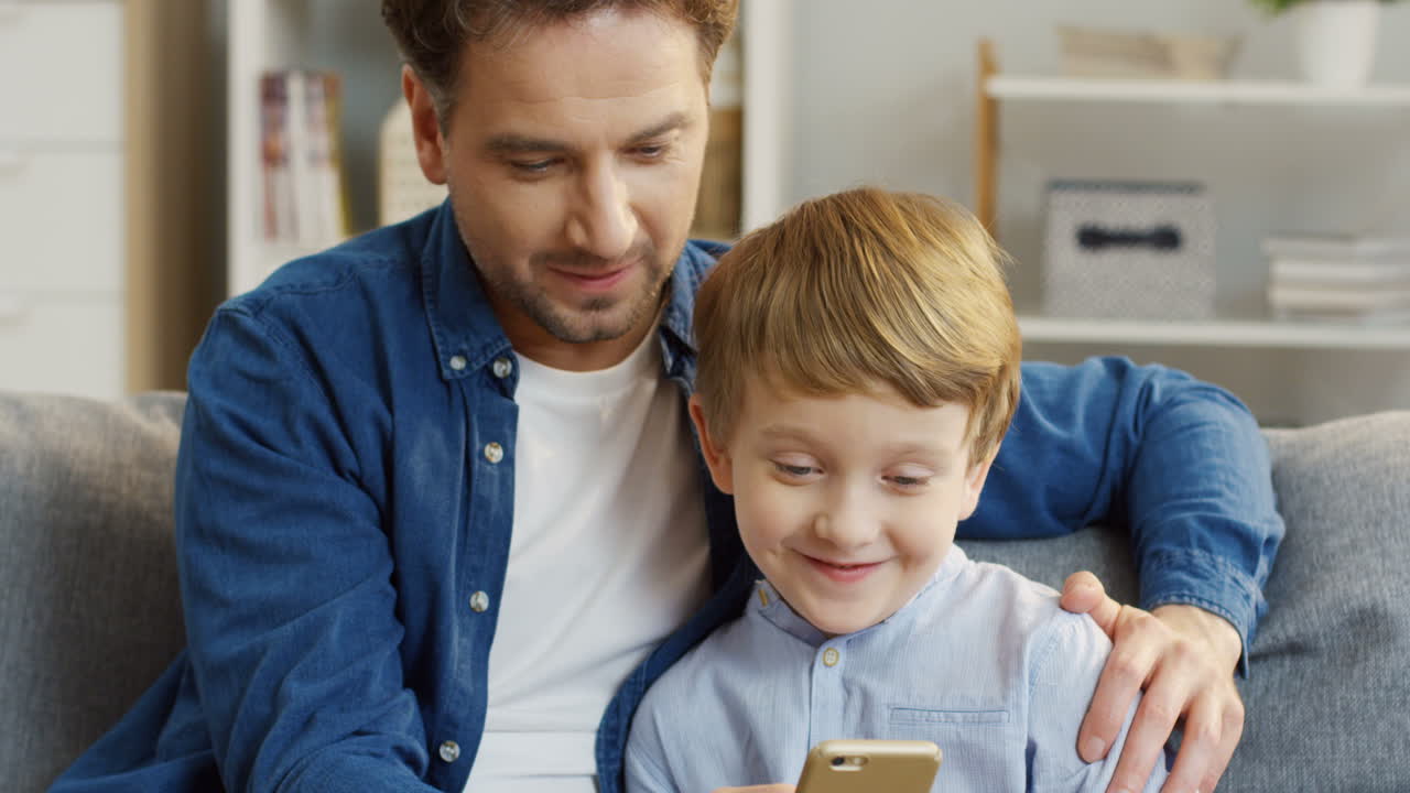 lindo niño rubio sentado al lado de su joven y apuesto padre, jugando un juego en el teléfono inteligente y sonriendo