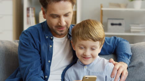 lindo niño rubio sentado al lado de su joven y apuesto padre, jugando un juego en el teléfono inteligente y sonriendo