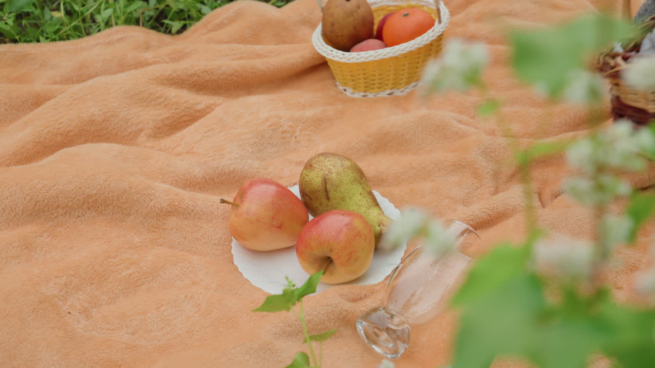 melocotones, peras y picnic sobre una manta naranja; cestas de mimbre y tarros de cristal sobre una tela suave; mano colocando una cesta pequeña; fondo de hierba; luz cálida del sol; ambiente rústico y acogedor; primer plano de fruta fresca y madura