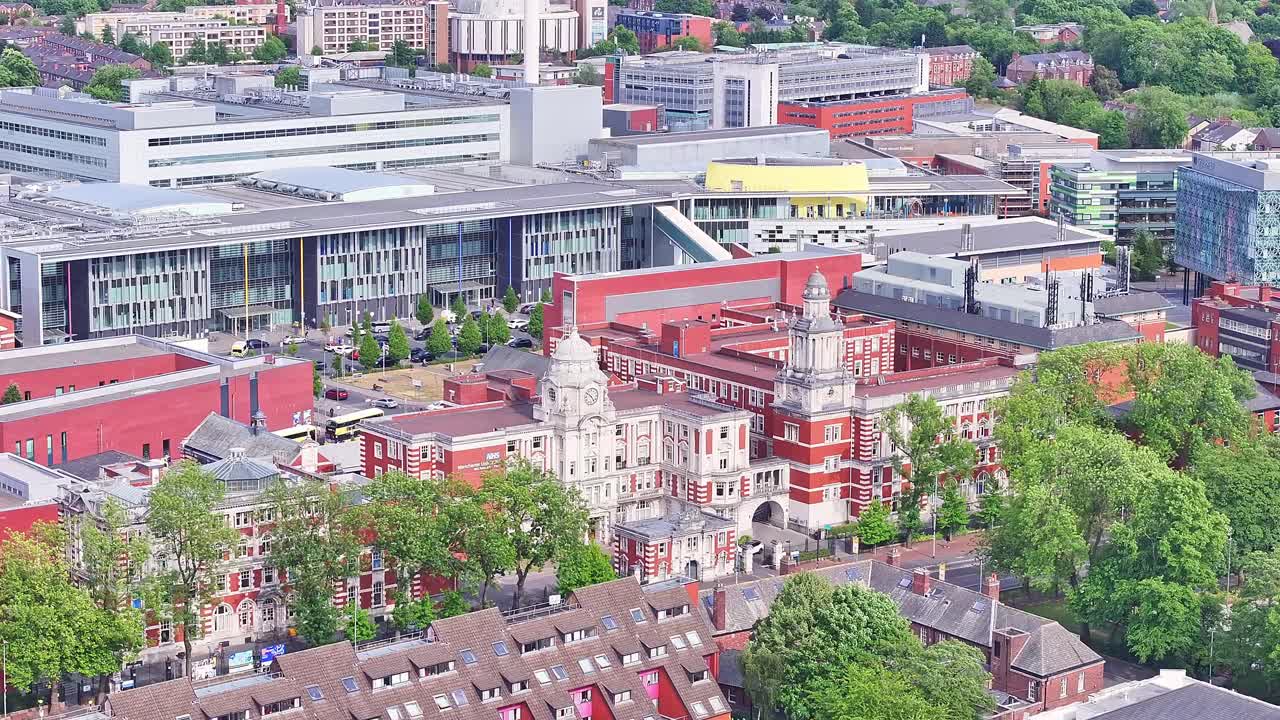 Aerial ascending shot from Dilworth Street showing University of Manchester campus with red-brick Victorian architecture, modern research buildings and green urban surroundings in Manchester, UK